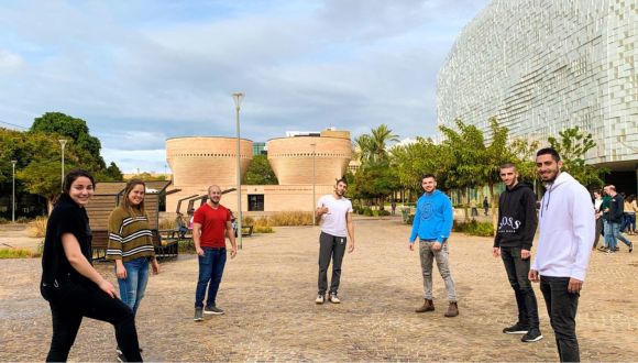 Seven students standing in a semi-circle on Tel Aviv University's campus, with the Cymbalista synagogue behind them and the Checkpoint building on the right