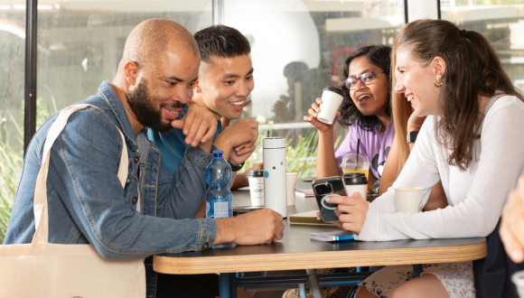 four students sitting at a table - two male students on the left and two female students on the right. They are smiling and are engaged in conversation 